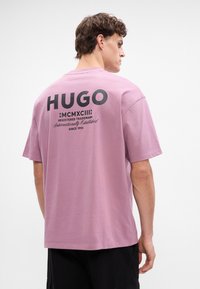 Ružový bavlnený tričko s veľkým čiernym logom "HUGO" na zadnej strane, s ďalším textom a rokom založenia nižšie. Krátke rukávy, okrúhly výstrih.