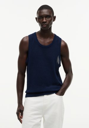 Mężczyzna ubrany w granatowy siateczkowy tank top bez rękawów i białe spodnie, stojący z jedną ręką w kieszeni na jednolitym tle.