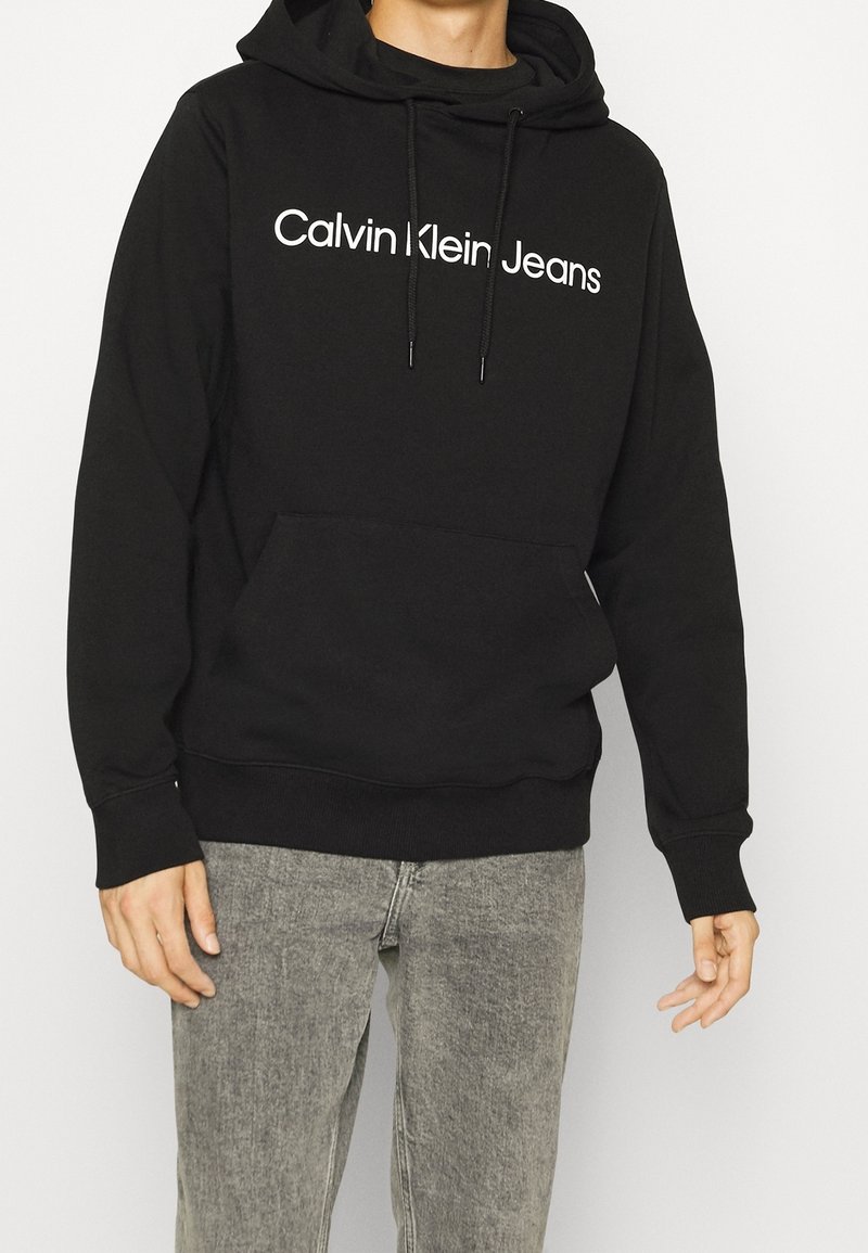Personne portant un sweat à capuche noir avec le texte "Calvin Klein Jeans" et un jean gris délavé, debout devant un fond blanc uni.