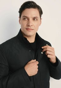 Czarna pikowana kurtka z wysokim kołnierzem; posiada zatrzaski i zamek błyskawiczny. Model poprawia kołnierz, nosząc czarną koszulę pod spodem.