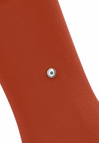 Chaussette couleur rouille en tissu texturé, présentant une étiquette circulaire en argent portant un logo, positionnée sur un fond blanc.