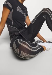 Svart träningsskjorta med Puma-logotyp, i kombination med svarta leggings med strukturerade mönster och vita sneakers med svarta detaljer.