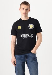 T-shirt en coton noir avec des graphiques circulaires en jaune et blanc. Le texte comprend "SUNNIES E.C." et "Lyle & Scott", avec une silhouette ajustée.