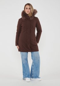 Cappotto isolato marrone con cappuccio foderato in pelliccia, dotato di tasche con zip e una linea lunga e dritta. Abbinato a jeans svasati blu chiaro e sneakers bianche.