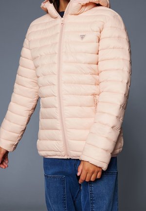 Enfant portant une veste matelassée rose clair avec capuche et poches zippées, associée à un jean bleu sur un fond gris uni.