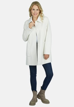 Femme aux cheveux blonds portant un manteau blanc boutonné, un jean skinny foncé et des bottines beiges, debout devant un fond uni.