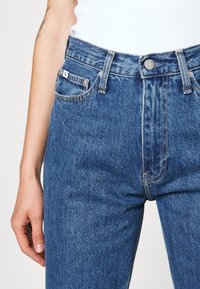 Hoge spijkerbroek van blauw denim met vijf zakken, een knoopsluiting en zichtbare stiksels, die een gestructureerde stofafwerking tonen.