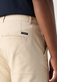 Beige broek van katoen met een gladde structuur, voorzien van een achterzak en een gestikt merklabel in zwart.