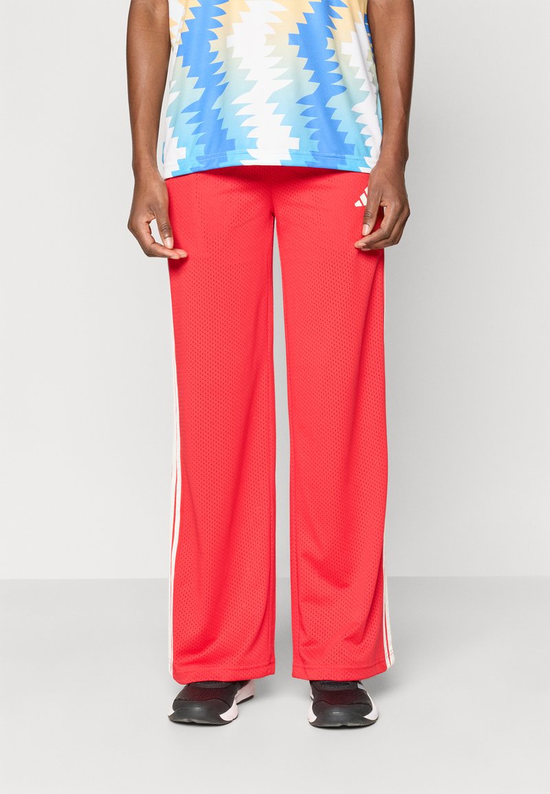 Osoba ubrana w jasnoczerwone szerokie spodnie dresowe z perforacją i białymi paskami po bokach, w czarnych sneakersach, stojąca na tle jednolitym.