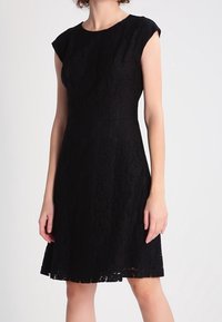Schwarzes Spitzenkleid mit kurzen Ärmeln, tailliertem Oberteil und ausgestellt Rock. Highlight ist das filigrane Spitzenmuster und der gewellte Saum.