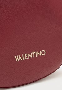Punainen nahkalaukku, jossa on teksturoitu pinta ja kultainen "VALENTINO"-logo modernilla fontilla. Yksinkertainen, pyöreä muotoilu, jossa on sileät reunat.