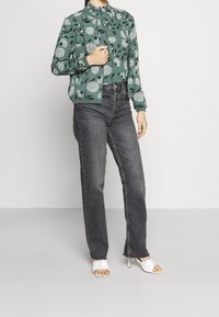 Grün gefütterte Bomberjacke mit floralem Muster und hohem Kragen, kombiniert mit hoch taillierten dunklen Jeans und weißen Absatzsandalen.