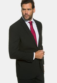 Veste noire avec revers crantés, assortie à une chemise blanche et une cravate texturée rose foncé. L'homme se tient debout, confiant, une main sur la veste.