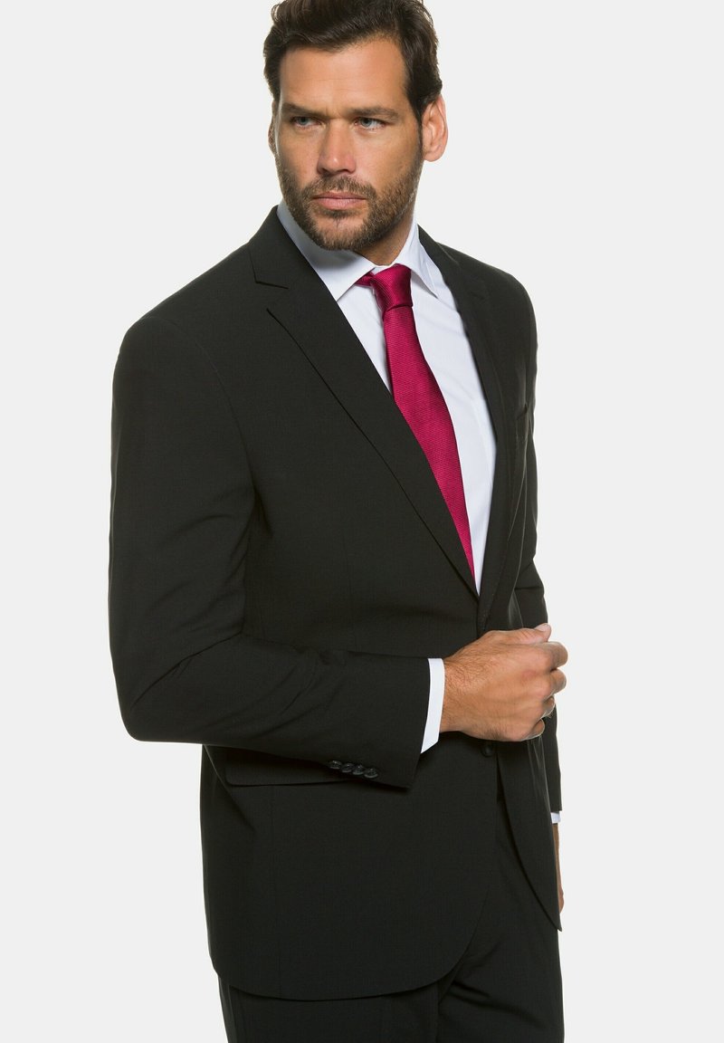 Veste noire avec revers crantés, assortie à une chemise blanche et une cravate texturée rose foncé. L'homme se tient debout, confiant, une main sur la veste.