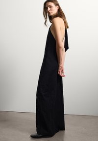 Un vestito nero senza maniche con una scollatura profonda, realizzato in un tessuto texturizzato e con una vestibilità morbida che arriva fino alle caviglie. In primo piano sono visibili un paio di scarpe basse.