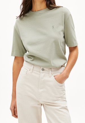 Frau trägt ein hellgrünes T-Shirt mit kurzen Ärmeln, das in eine hoch taillierte off-white Hose gesteckt ist, mit einer Hand in der Tasche.