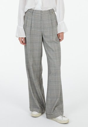 Woman wearing gray plaid wide-leg pants, white blouse with flared cuffs, and white sneakers, standing against a plain background.