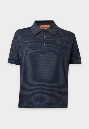 Ung mand står med den ene hånd i lommen iført mørk, stribet polo, vide lyseblå jeans og sorte sko på hvid baggrund.