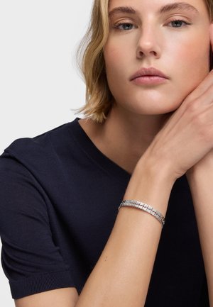 Mujer con cabello rubio corto que lleva una blusa azul marino, apoyando su rostro en la mano y mostrando una pulsera plateada con un diseño en su muñeca.