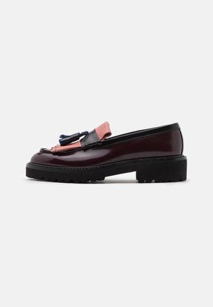 Dunkelburgunderfarbener Leder-Slipper mit schwarzer Sohle, rosa Lederfransen auf der Zunge und schwarzem sowie navyblauem Quastdetail vor weißem Hintergrund.