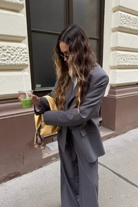 Pelēku svītroto uzvalku ar plašiem biksēm un pielāgotu žaketi; tur zaļu dzērienu un dzeltenu somu ar piekariņu.