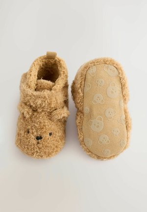 Chaussons pour tout-petits en peluche tan avec un design de visage d'ours, texture douce et une semelle en caoutchouc à motifs comportant de petites têtes d'ours.