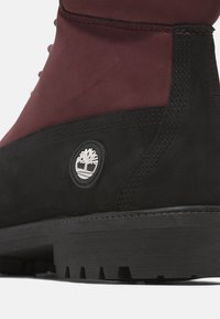 Bottine noire et rouge foncé, en suede avec une finition texturée, dotée d'un logo circulaire, de coutures visibles et d'une semelle extérieure robuste.