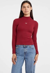 Egy nő fazonra szabott, piros, hosszú ujjú, álló nyakú felsőt visel, amin a "Guess Jeans" logó látható, valamint magas derekú, világoskék farmert, és egy fehér háttér előtt áll.