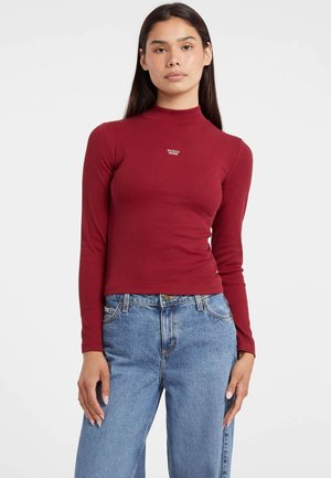 Naine kannab hästi istuvat punast pikavarrukaga toppi kõrge kaelusega, millel on "Guess Jeans" logo, ja kõrge vöökoha ning helesiniseid teksaseid, seistes valge tausta ees.