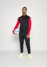 Schwarzes und rotes Sportoberteil mit einem halben Reißverschluss und Logo, kombiniert mit schwarzen Hosen mit seitlichen Knöpfen und neongrünen Schuhen, während ein Fußball gehalten wird.