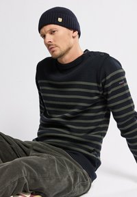 Homme assis portant un bonnet bleu marine avec un logo d'ancre, un pull rayé noir et olive avec des boutons à l'épaule, et un pantalon en velours côtelé gris.