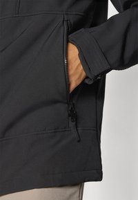 Crna softshell jakna s patentnim zatvaračem na bočnoj strani. Značajke uključuju šivane šavove i blago teksturiranu tkaninu.