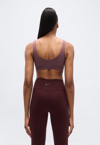 Reggiseno sportivo in viola spento e leggings a vita alta in bordeaux, con logo grigio. Tessuto liscio e aderente con spalline larghe.