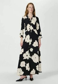 Vestido maxi preto com grande padrão floral branco, design com botões à frente, mangas compridas e um tecido suave. Usado com sapatos de salto pontiagudo pretos.