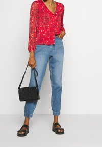 Rød blomstret bluse med V-udskæring, lange ærmer, parret med lyseblå jeans. Holder en lille vævet sort håndtaske og bærer sorte sandaler.