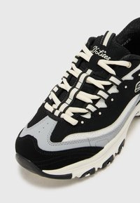 Black and grey sneaker with a mesh upper, white laces, and a thick sole. Features curved accents and subtle logo detailing on the tongue.