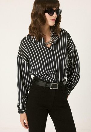 Femeie care poartă o bluză cu dungi verticale alb-negru, pantaloni negri cu curea, ochelari de soare închiși la culoare și un colier gros de argint.