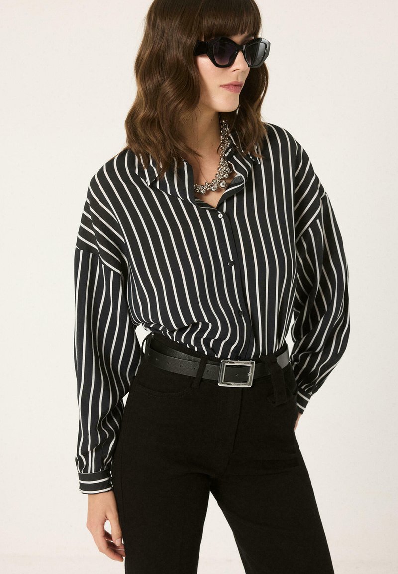 Femme portant un chemisier à rayures verticales noir et blanc, un pantalon noir avec ceinture, des lunettes de soleil foncées et un collier argenté épais.