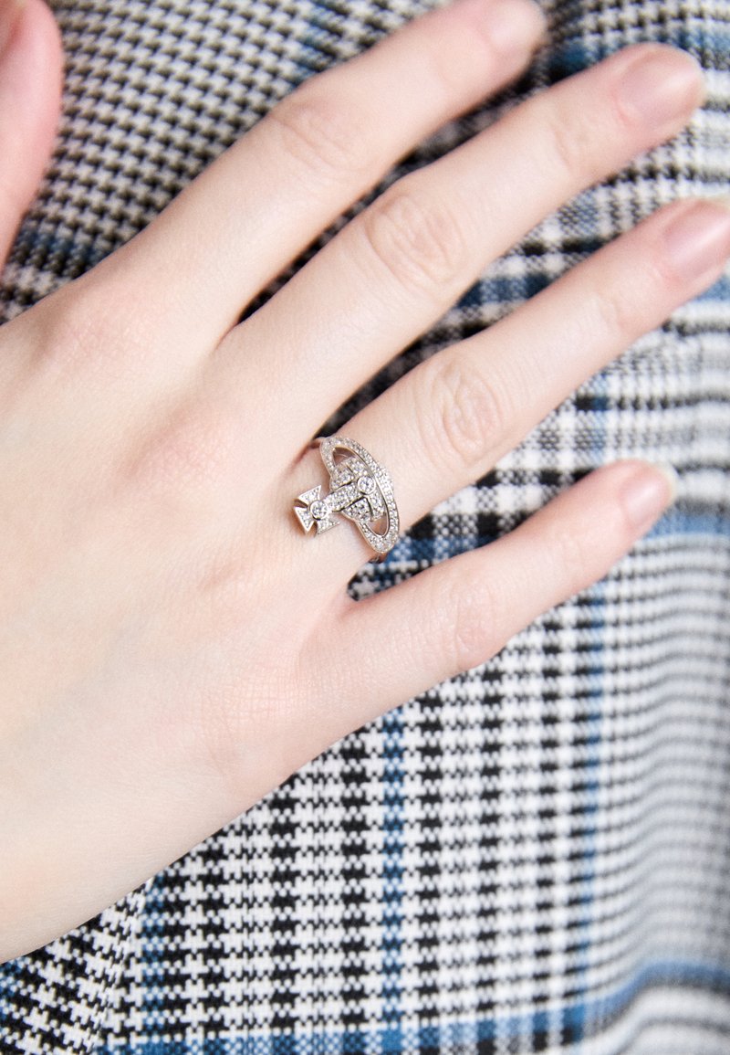 Anillo de plata con un diseño de lazo, incrustado con pequeñas piedras transparentes, exhibido en una mano sobre un fondo textil a cuadros.
