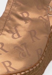 Κοντινό πλάνο από μπεζ ύφασμα με επαναλαμβανόμενα καφέ γράμματα "R", ραμμένο σε υφαντή σόλα από φυσικές ίνες, που προτείνει ένα casual εσπαντρίγια.