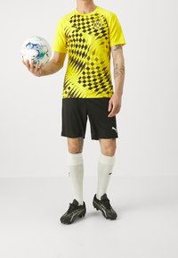 Camiseta de fútbol con patrón amarillo y negro, pantalones cortos negros, calcetas blancas y botas negras; sosteniendo un balón de fútbol. Diseño texturizado y logotipo visibles.
