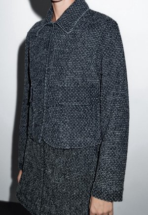 Person wearing a textured dark gray wool jacket with a large front pocket and collar against a plain white background.