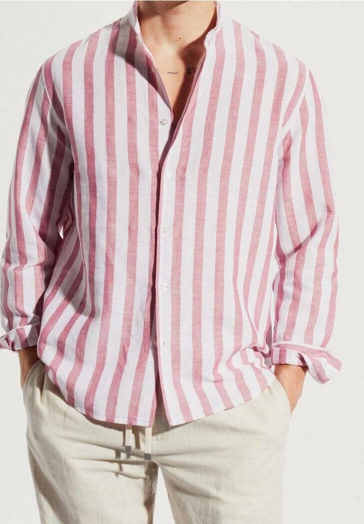 Homme portant une chemise à manches longues, boutonnée, rose et blanche à rayures verticales, pantalon beige clair, mains dans les poches, debout devant un fond uni.