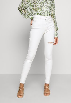 Vrouw die witte skinny jeans met hoge taille draagt, met een kleine scheur op de dij, beige sandalen met bandjes en een groen gebloemde blouse.