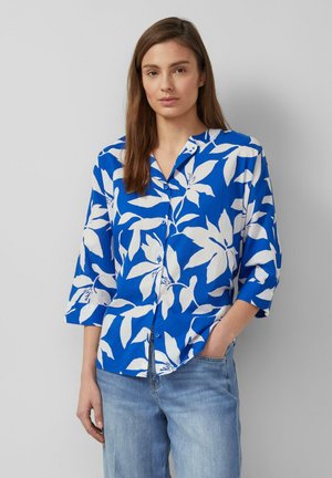 Frau mit langen braunen Haaren, die ein blaues Hemd mit großen weißen Blumenmustern und hellblaue Jeans trägt, steht vor einem grauen Hintergrund.