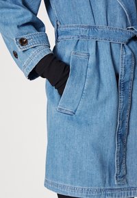 Modrý džínový kabát s páskem v pase, zapínáním na knoflíky na manžetách a bočními kapsami. Látka má světlé vyprání a hladkou texturu.