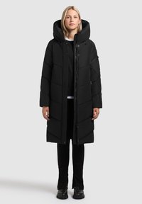 Abrigo de plumas negro con cuello alto, con cierre de cremallera en la parte frontal, textura acolchada y mangas largas. Lleva puesto sobre pantalones negros y botas.