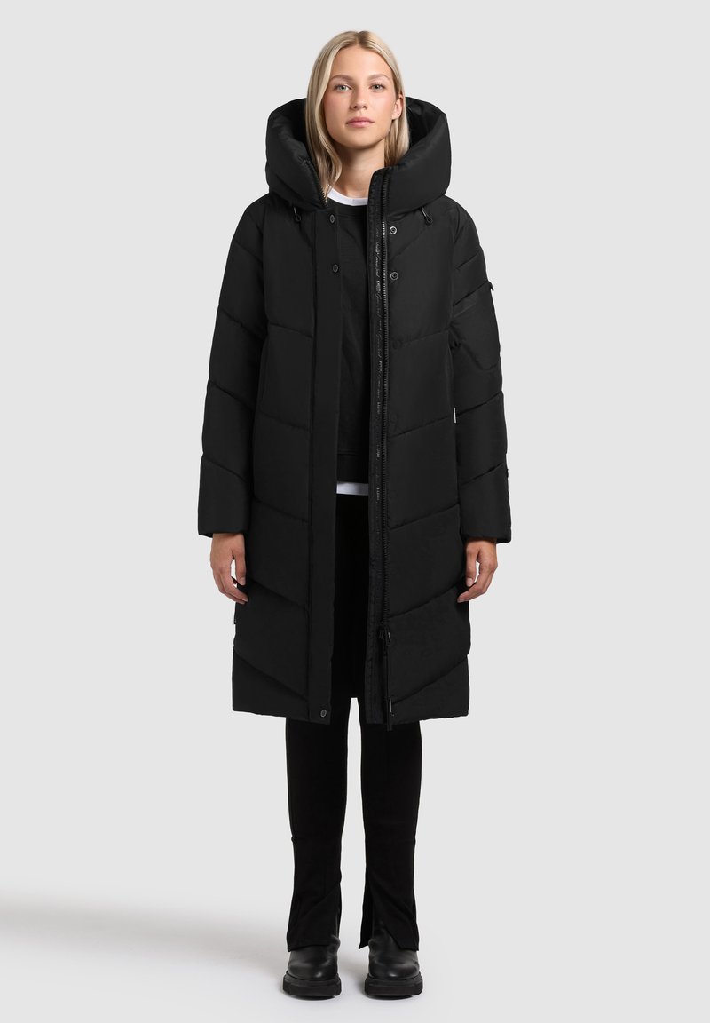 Abrigo de plumas negro con cuello alto, con cierre de cremallera en la parte frontal, textura acolchada y mangas largas. Lleva puesto sobre pantalones negros y botas.