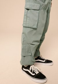 Grüne Cargo-Hosen mit großen Seitentaschen, umgeschlagenen Säumen und einer leichten Textur, kombiniert mit schwarzen Vans Sneakers mit weißen Akzenten.