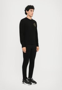 Μαύρη μπλούζα με στρογγυλή λαιμόκοψη και ριγέ μανσέτες, με χρυσό κυκλικό λογότυπο στο στήθος, συνδυασμένη με μαύρο παντελόνι και μαύρα αθλητικά παπούτσια.